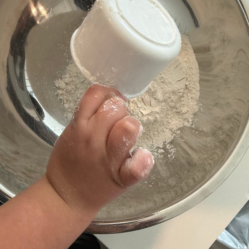 A small hand tipping a cup of flour into a steel mixing bowl